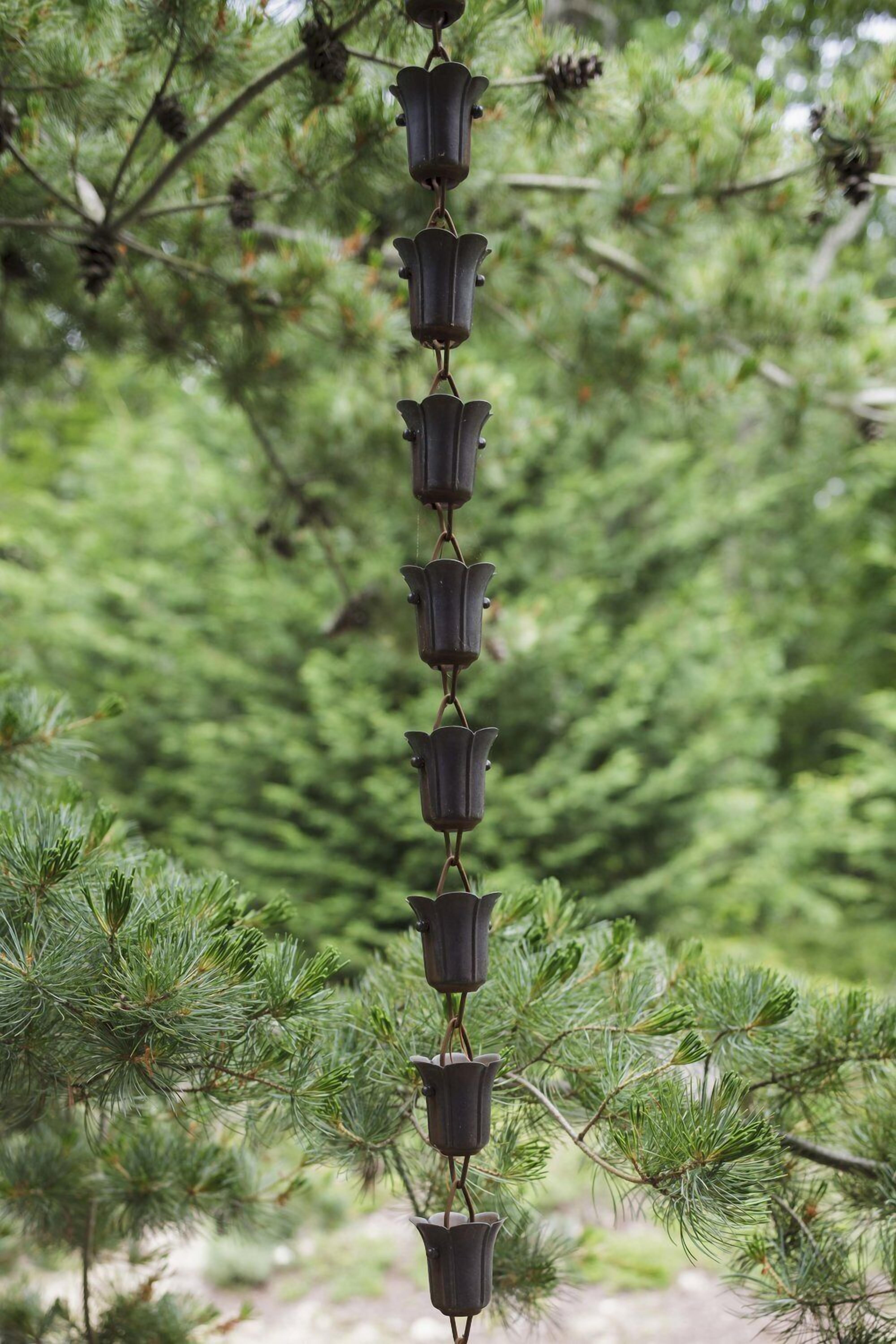A vertical arrangement of metallic bell-shaped objects hanging from a chain, set against a backdrop of dense green foliage.