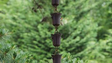 A vertical arrangement of metallic bell-shaped objects hanging from a chain, set against a backdrop of dense green foliage.