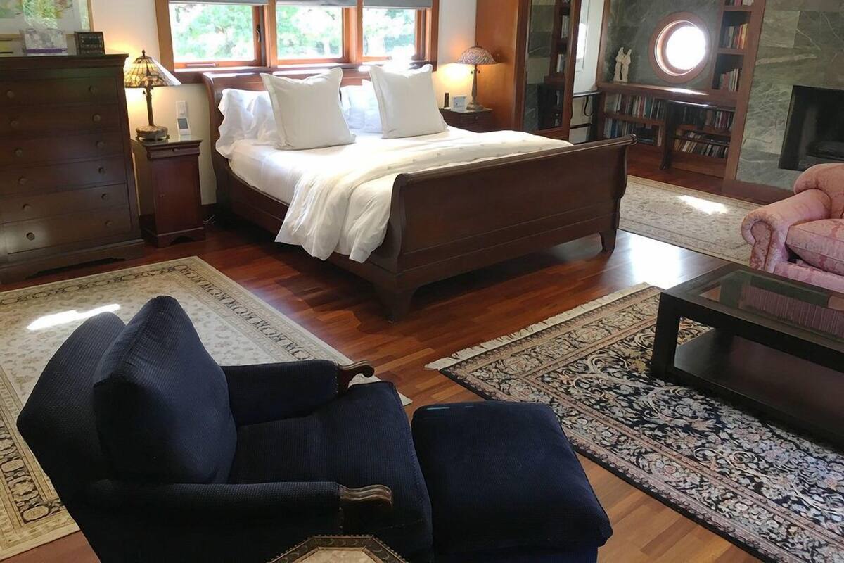 A bedroom with a large bed, two bedside tables with lamps, a wooden dresser, a chair, a coffee table, and a fireplace.