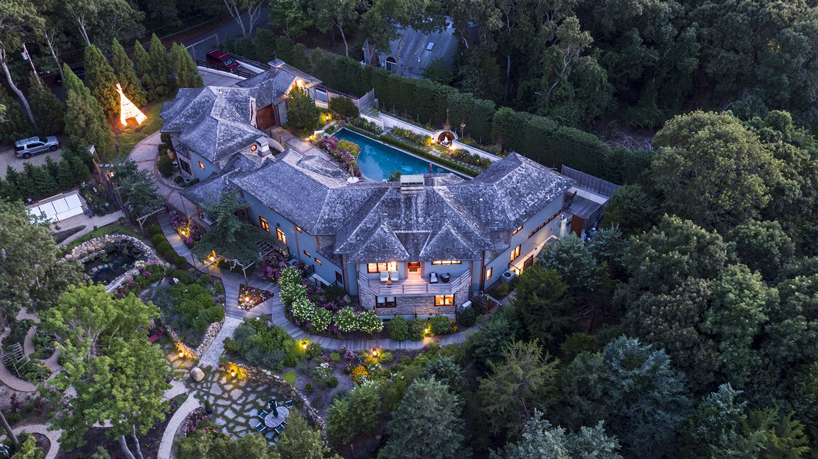 A large, two-story house with a swimming pool, surrounded by dense trees and landscaped gardens.