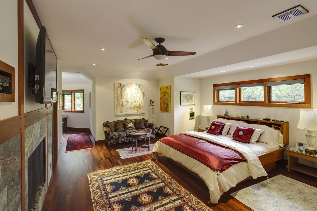 A spacious bedroom with a large bed, a ceiling fan, a fireplace, and a view of the outdoors through a window.