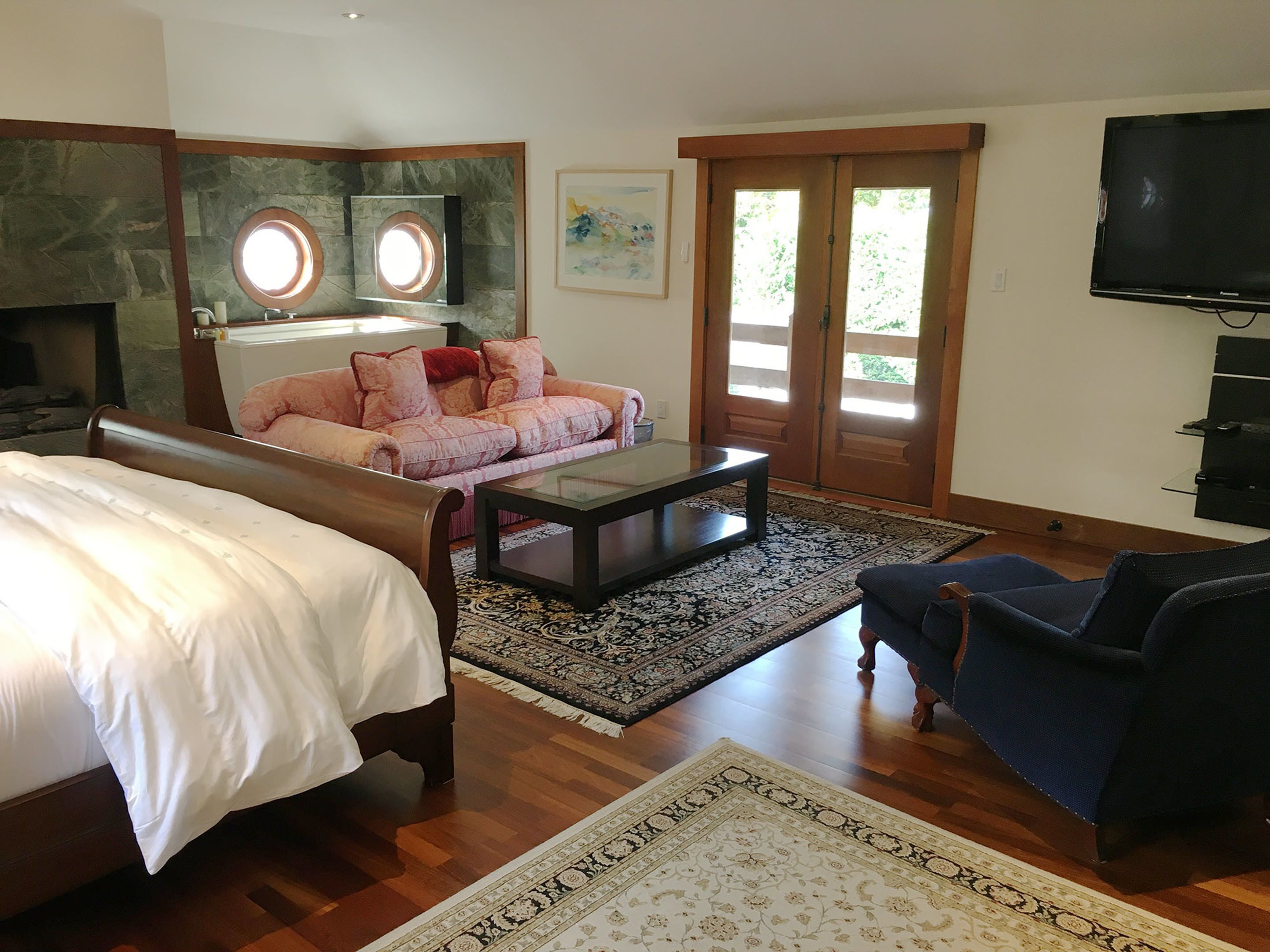 A spacious living room with a large bed, a sofa, a coffee table, and a TV. There is a fireplace and a large window with a view of greenery.