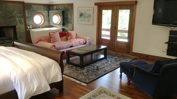 A spacious living room with a large bed, a sofa, a coffee table, and a TV. There is a fireplace and a large window with a view of greenery.