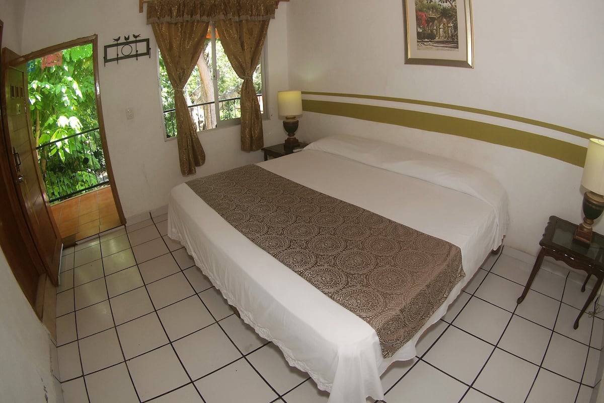 Um quarto com cama, cortinas, um quadro na parede, abajur e janela com vista para o verde.