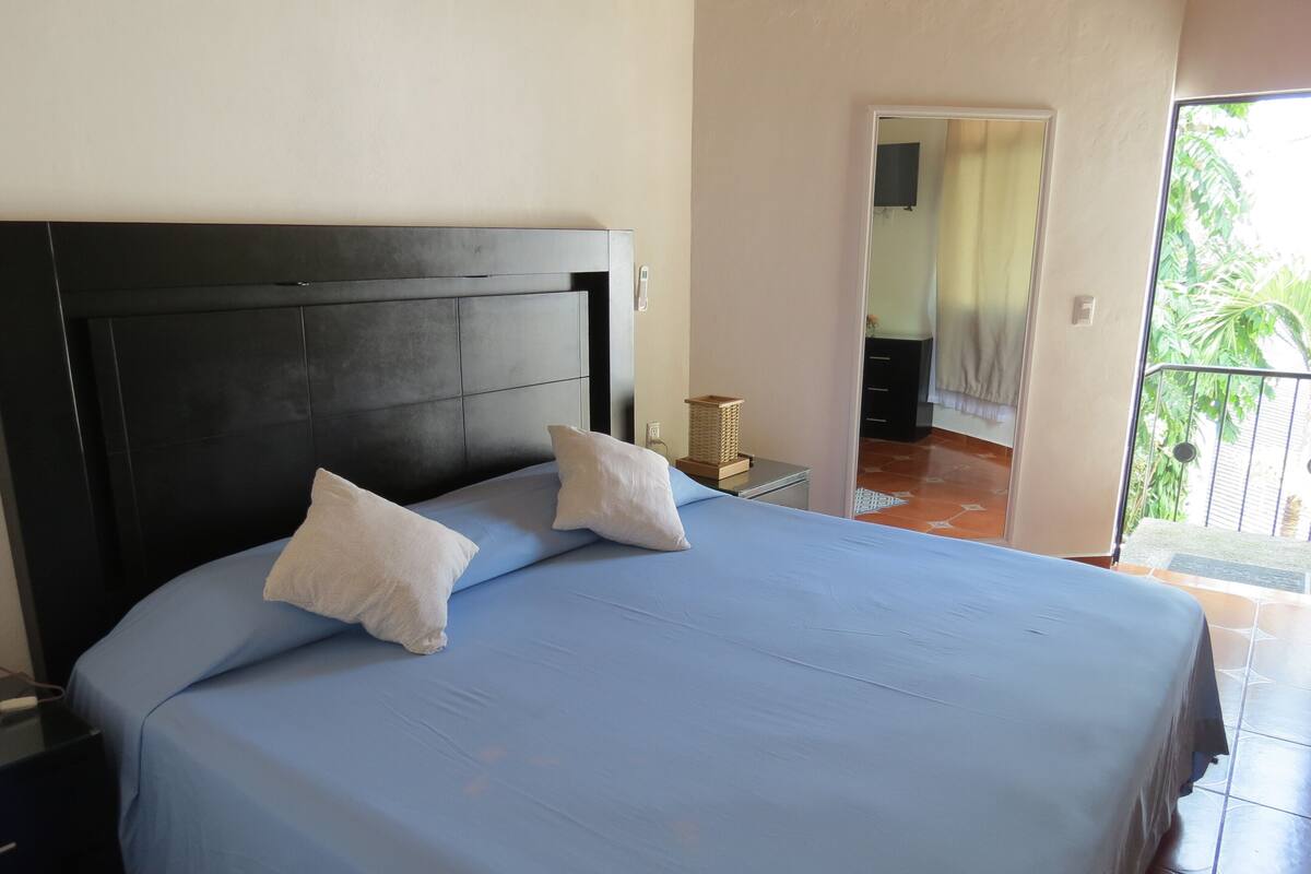 Quarto com uma cama grande, duas almofadas, uma mesa de cabeceira e janela com vista para o verde.