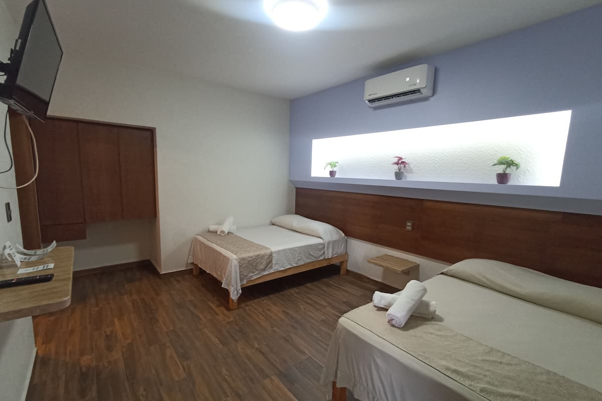 Quarto de hotel com duas camas de solteiro, piso de madeira, televisão, ar condicionado e janela com plantas.