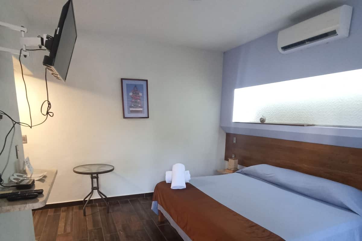 Quarto pequeno e limpo com uma cama, uma mesinha, televisão fixada na parede e um quadro enquadrado na parede.