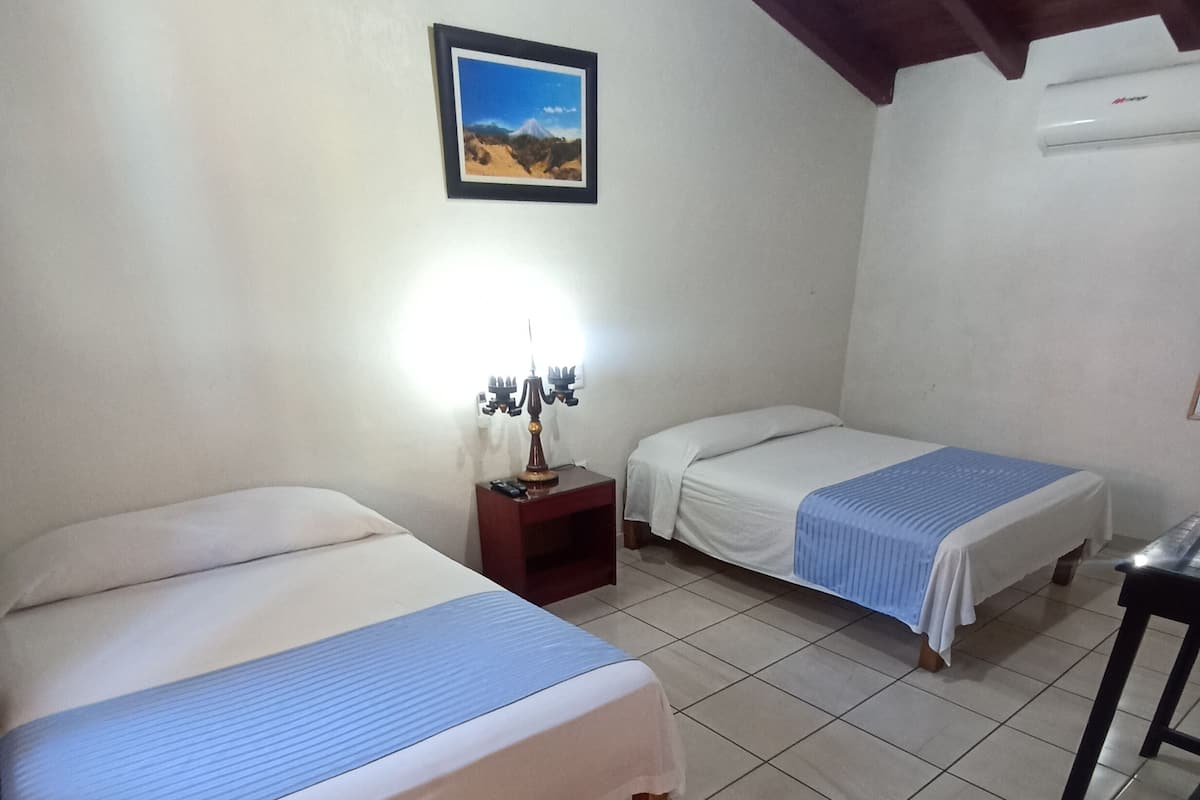 Quarto de hotel com duas camas, uma mesinha de cabeceira com luminária, um quadro na parede e um ar-condicionado.