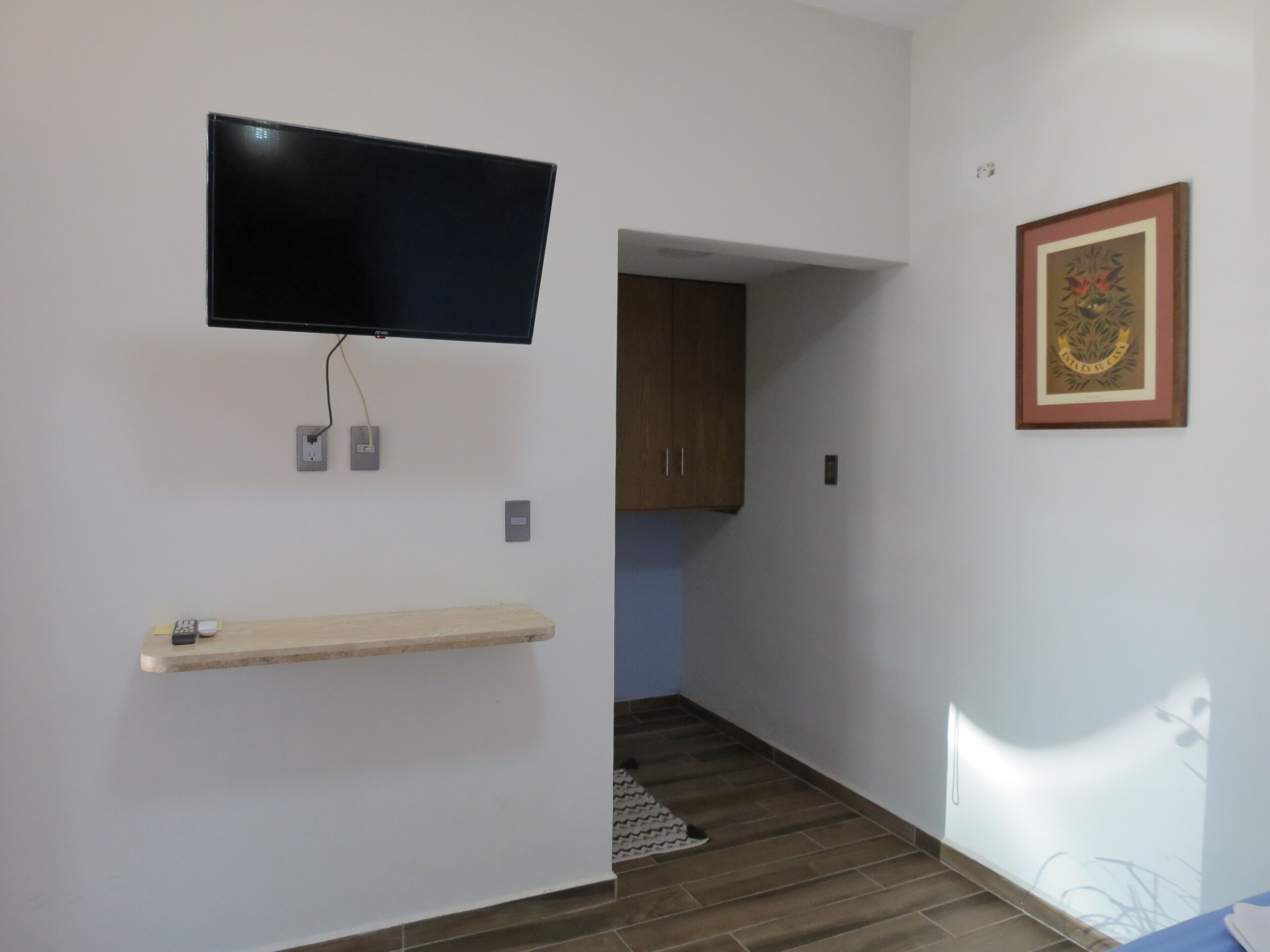 Quarto com televisão na parede, prateleira de madeira, quadro emoldurado e porta de acesso a outro espaço.