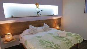 Una cama bien hecha con estampado de hojas verdes, dos lámparas de noche, un espejo sobre la cama y una planta pequeña sobre el espejo.