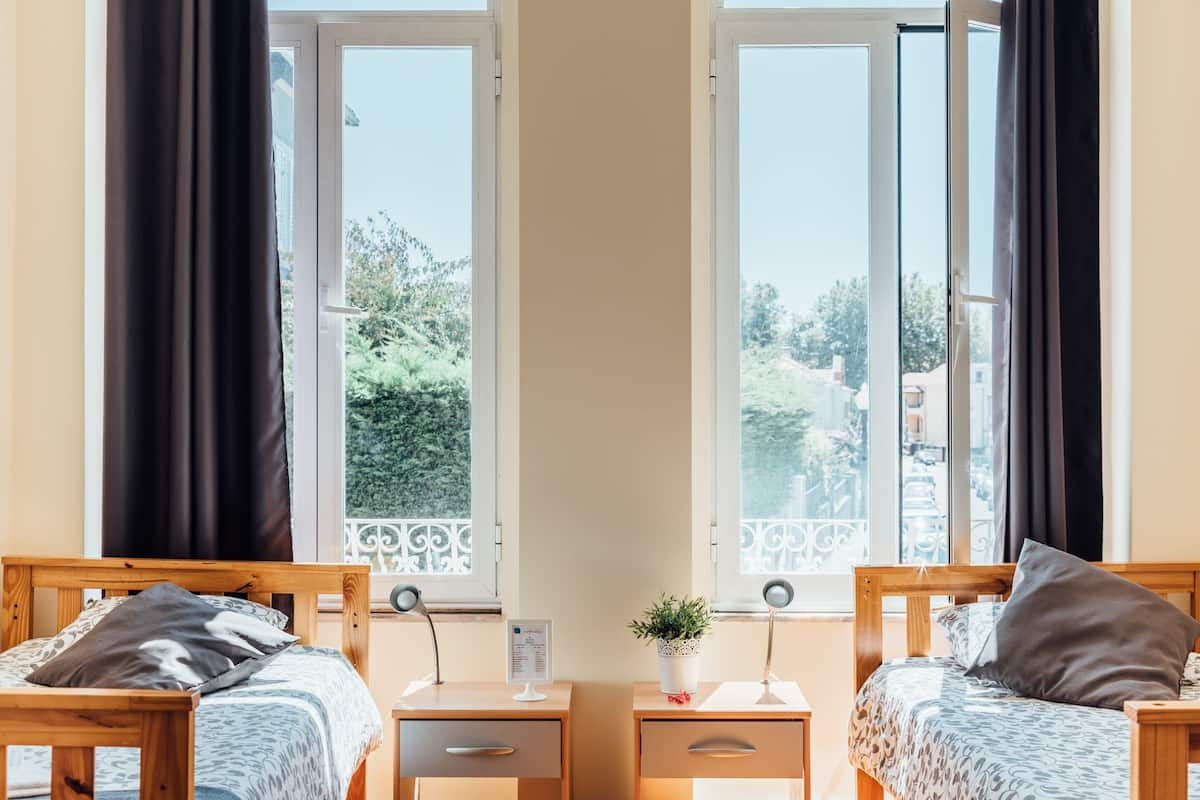 A room with two wooden beds, a small desk, and a window with a view of greenery and buildings.