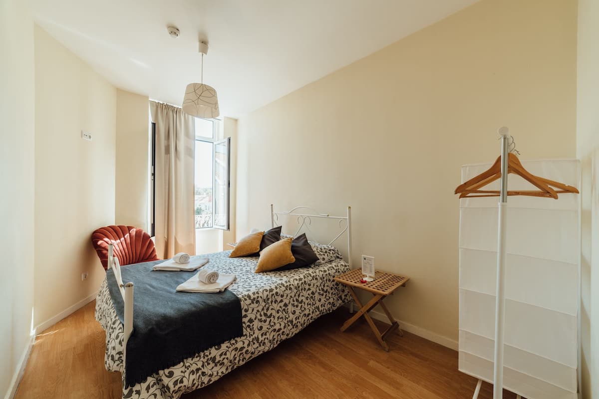 A bedroom with a bed, a chair, a small table, a hanging clothes rack, and a window with curtains.