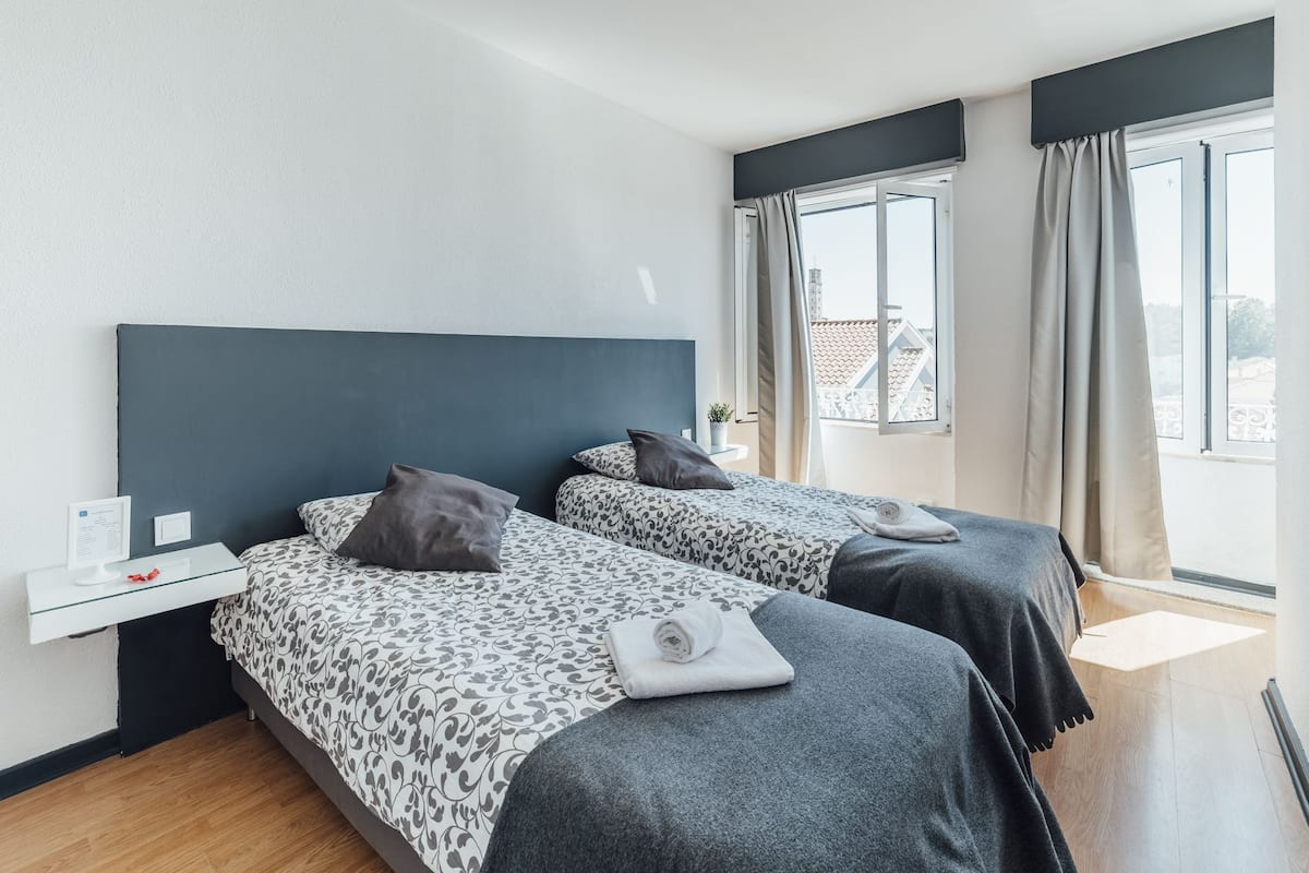 A hotel room with two beds, a small bedside table, a window with curtains, and a view of buildings outside.