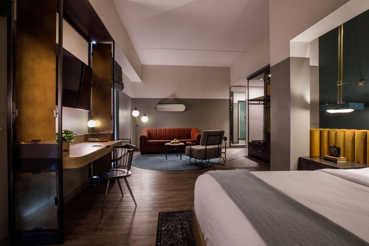 A modern hotel room with a large bed, a desk with a chair, a sofa, and a small table with a lamp.