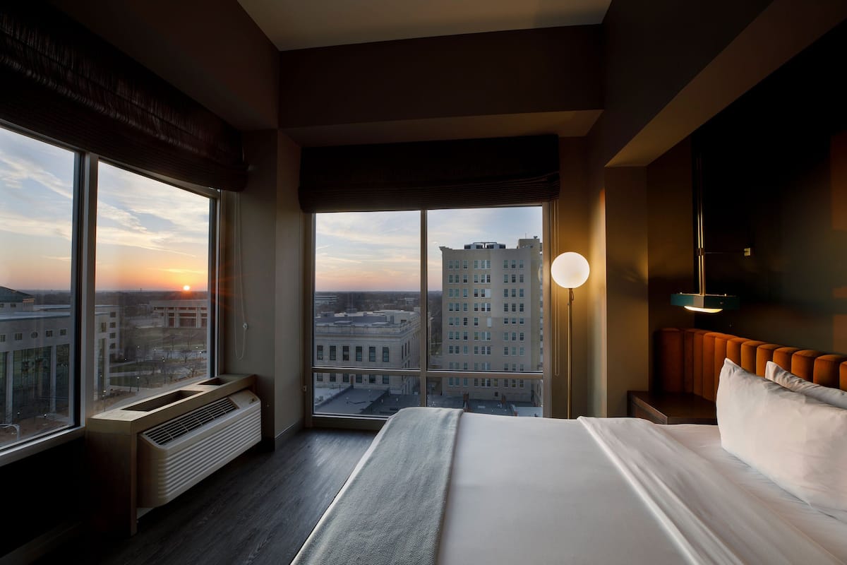 A hotel room with a large window, a bed, a heater, and a nightstand with a lamp.