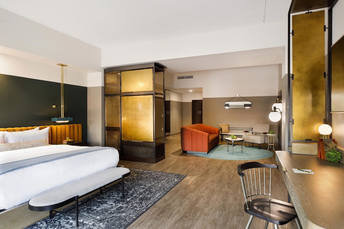 A modern hotel room with a large bed, a seating area with a bench and a chair, a small table, and a sofa.