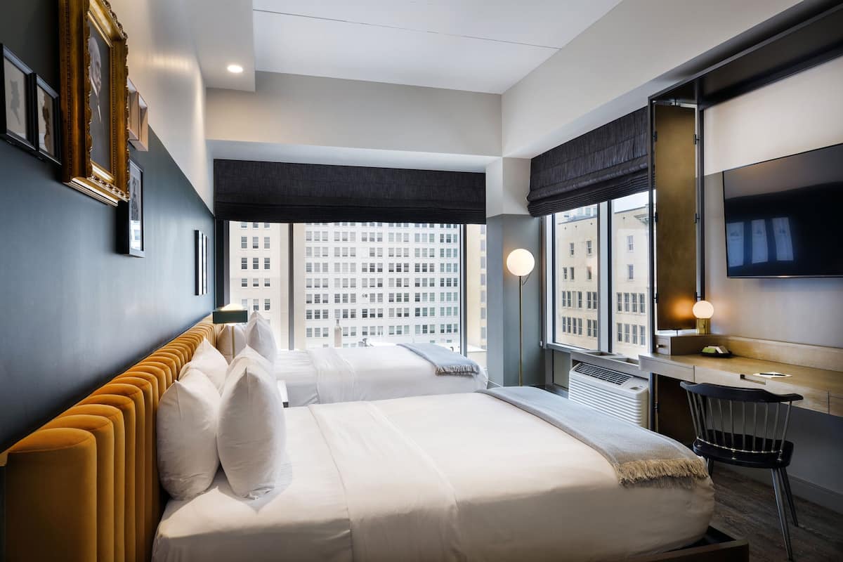 A modern hotel room with two beds, a desk, and a large window offering a city view.