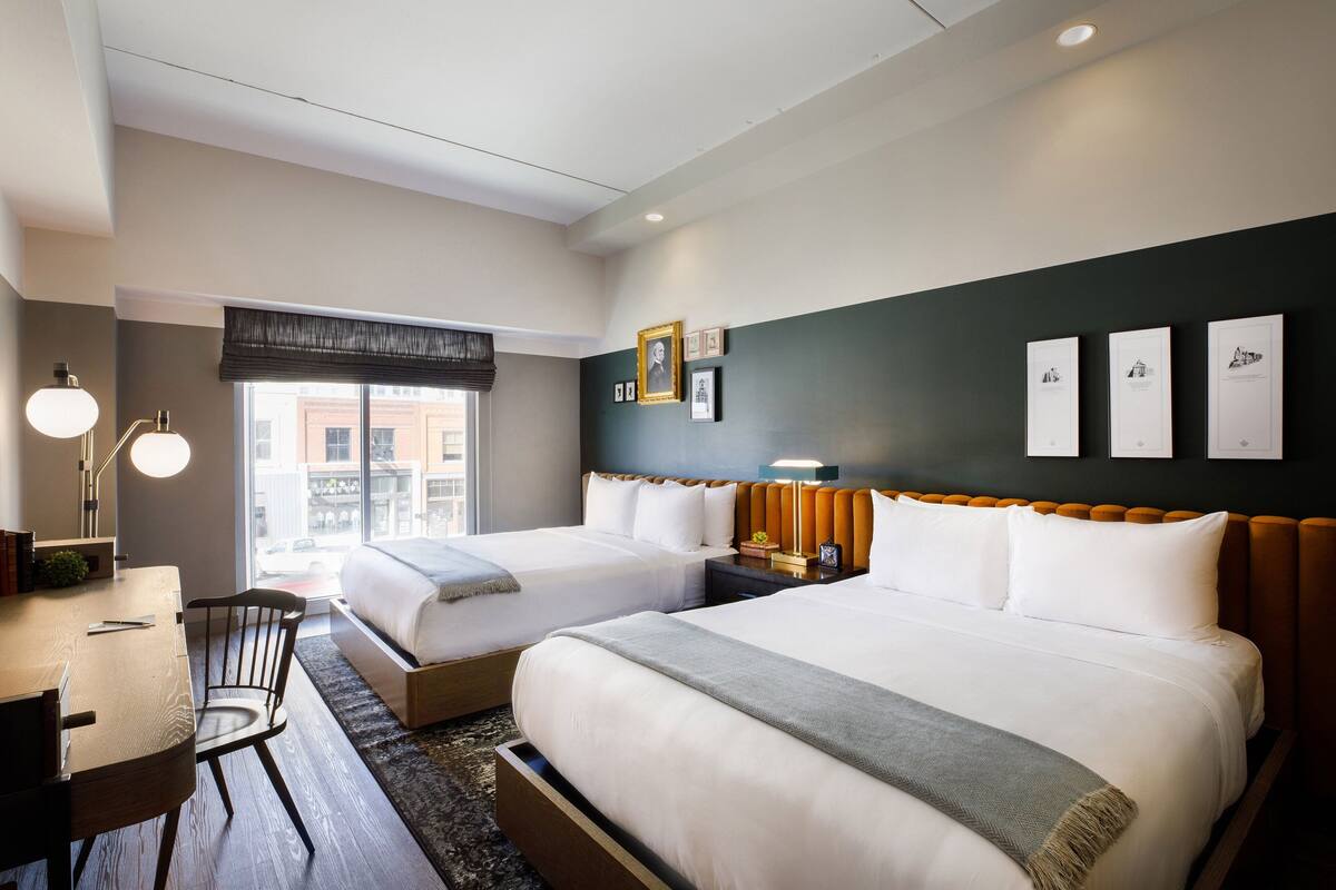 A hotel room with two beds, a desk, a chair, and a window with a view of buildings.