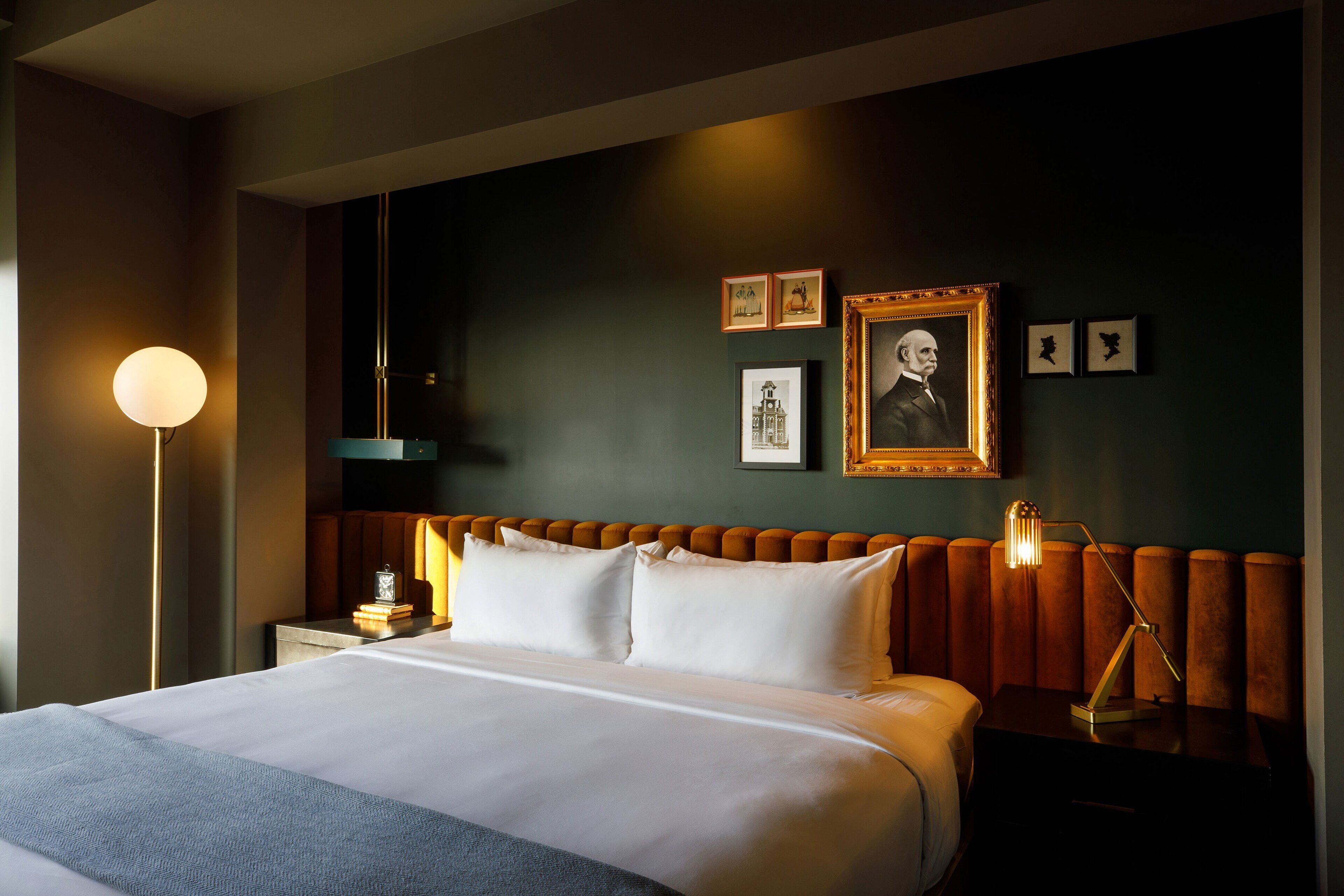 A neatly made bed with white linens, a bedside table with a lamp, and a framed portrait on the wall.