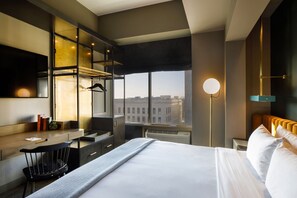 A modern hotel room with a large bed, a desk, and a chair. There is a large window with a view of buildings outside.