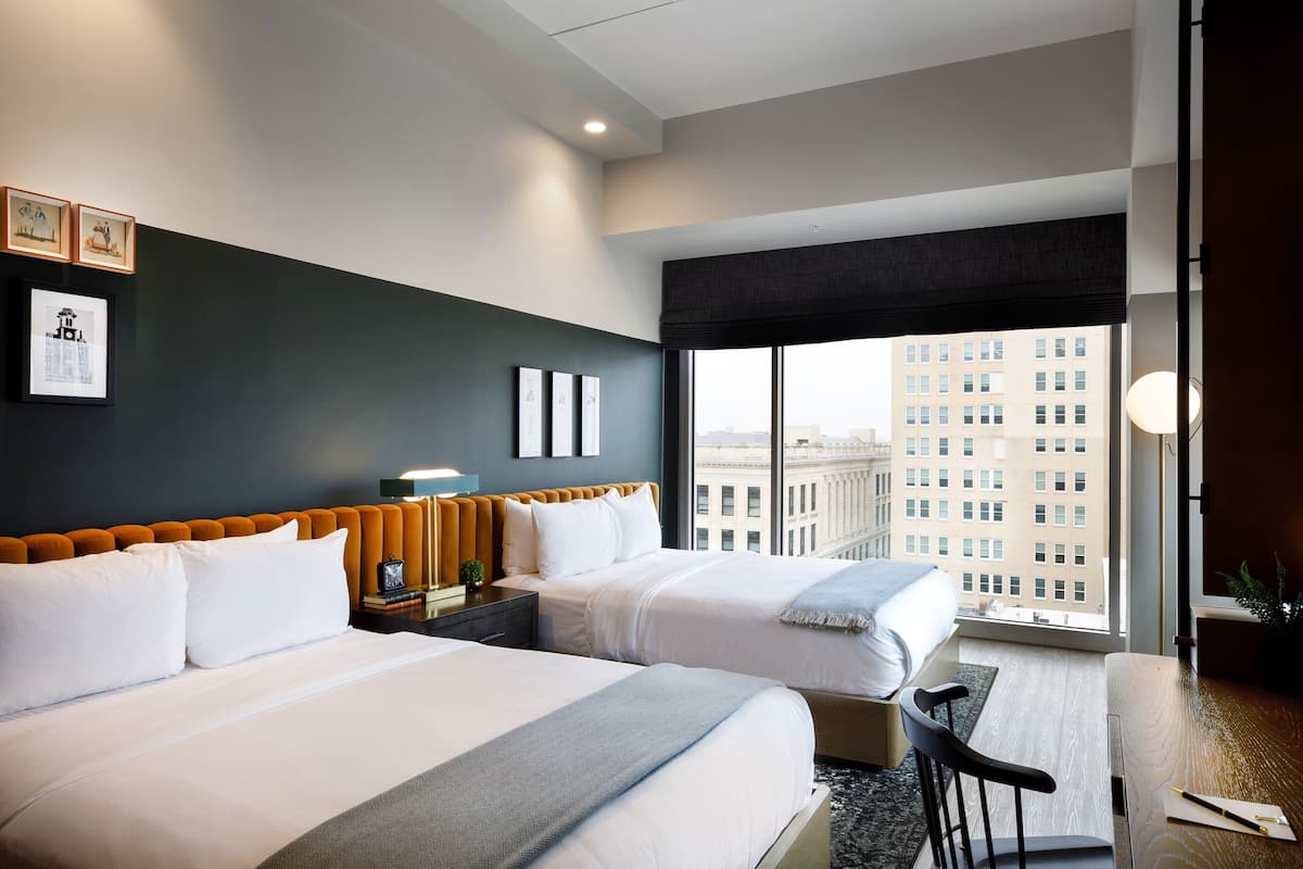 A hotel room with two beds, a desk, and a large window offering a city view.