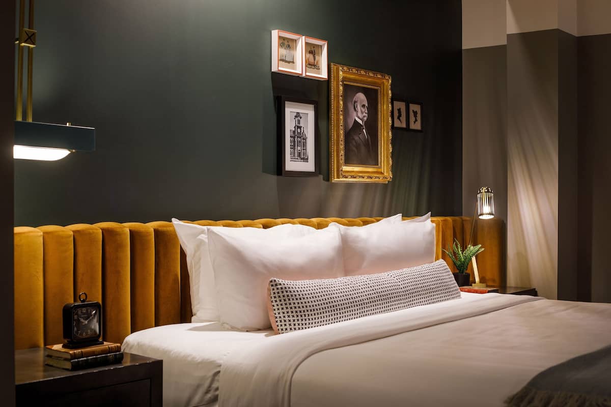 A neatly made bed with white linens, a mustard-colored headboard, and a bedside table with a clock and books.
