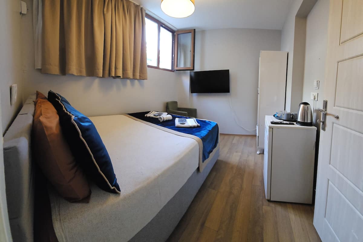 Quarto pequeno e compacto, com cama, escrivaninha, cadeira, televisão e cozinha compacta.