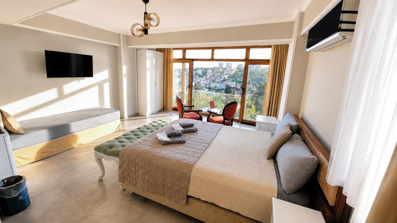 A modern hotel bedroom with a large bed, a seating area, a TV mounted on the wall, and a view of the city through large windows.