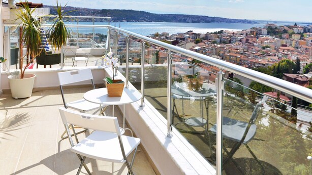 Un balcón con sillas y mesas blancas, plantas en macetas y una barandilla de cristal con vistas a la ciudad y al agua.
