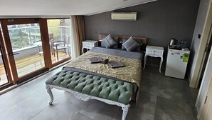 Un dormitorio con cama, mesitas de noche, un banco, un refrigerador y una ventana con vista a edificios.