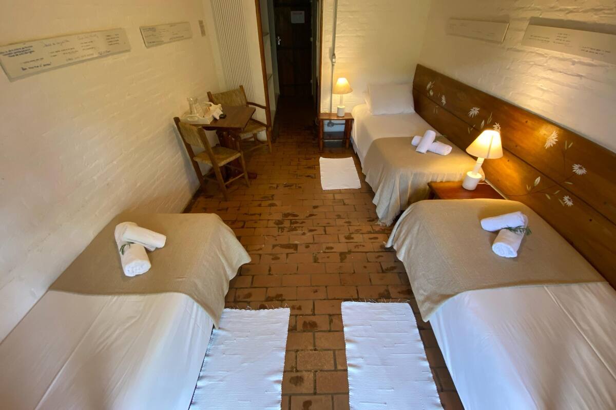 A hotel room with two single beds, a small table with chairs, a lamp, and a brick floor.