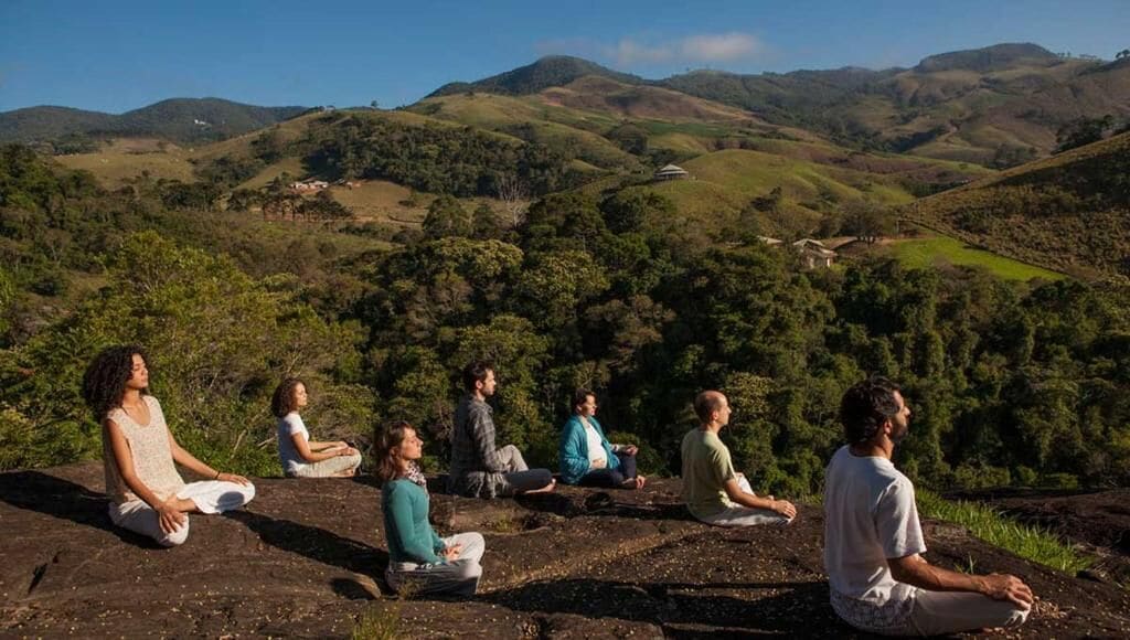 Eine Gruppe von Menschen meditiert auf einem Felsen mit herrlichem Blick auf sanfte Hügel und grüne Landschaft im Hintergrund.