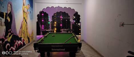 Una mesa de billar con superficie de fieltro verde, una vara de billar y una bola blanca. En la pared, a la izquierda de la mesa, hay un mural.