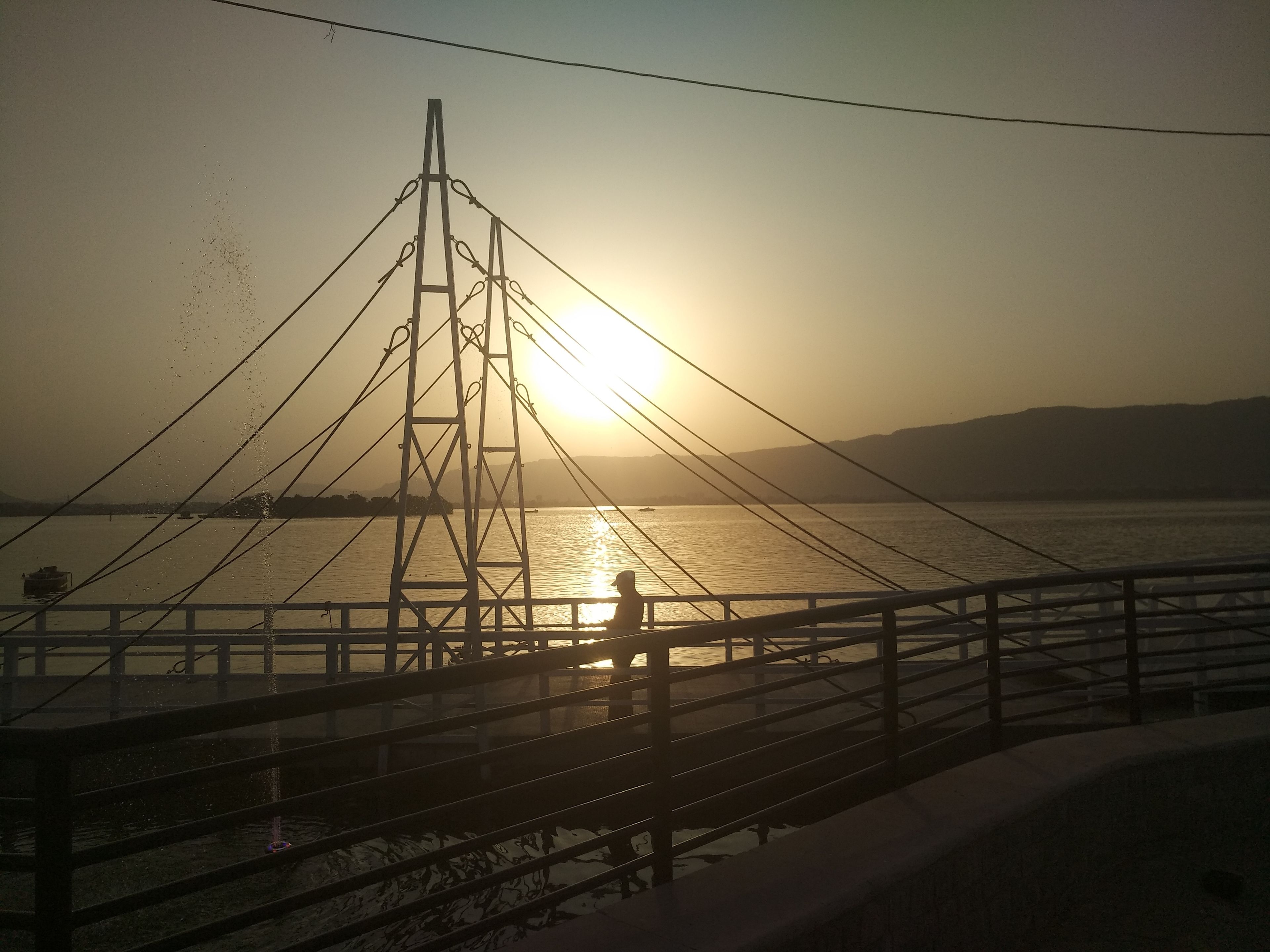 A bridge with a metal framework and cables, overlooking a body of water with a sunset in the background.