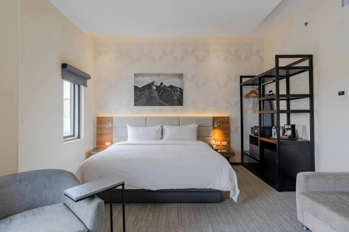 A modern hotel room with a large bed, a bedside table, a nightstand, a bookshelf, and a window.