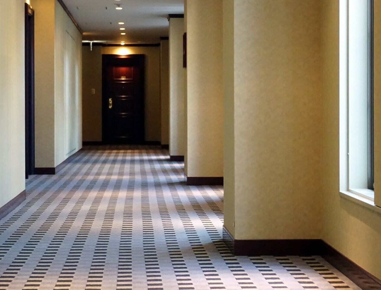 A hotel hallway with a checkered floor pattern, a door at the end, and walls with a light color.