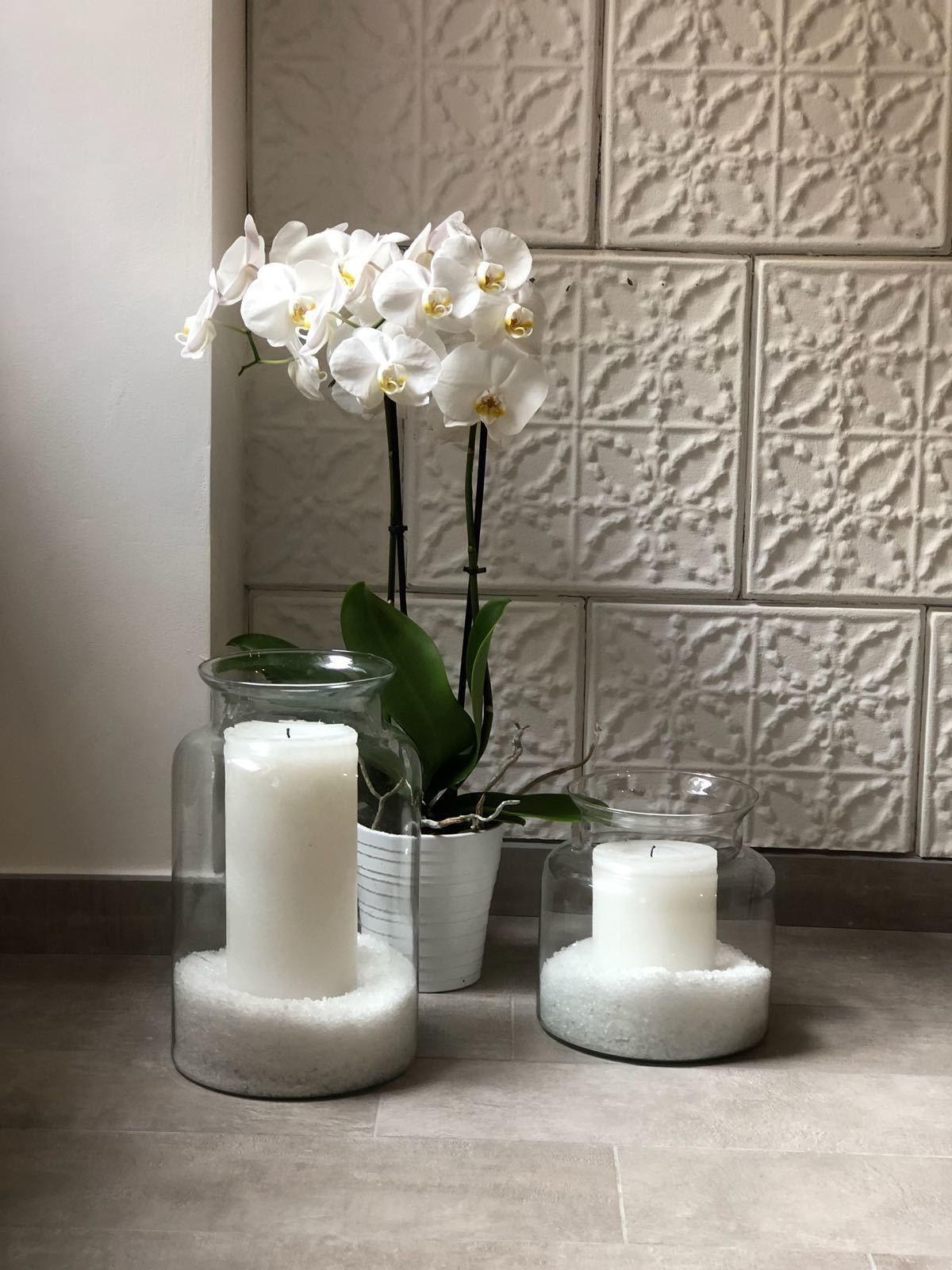 Three glass vases with white flowers and candles on a tiled floor.