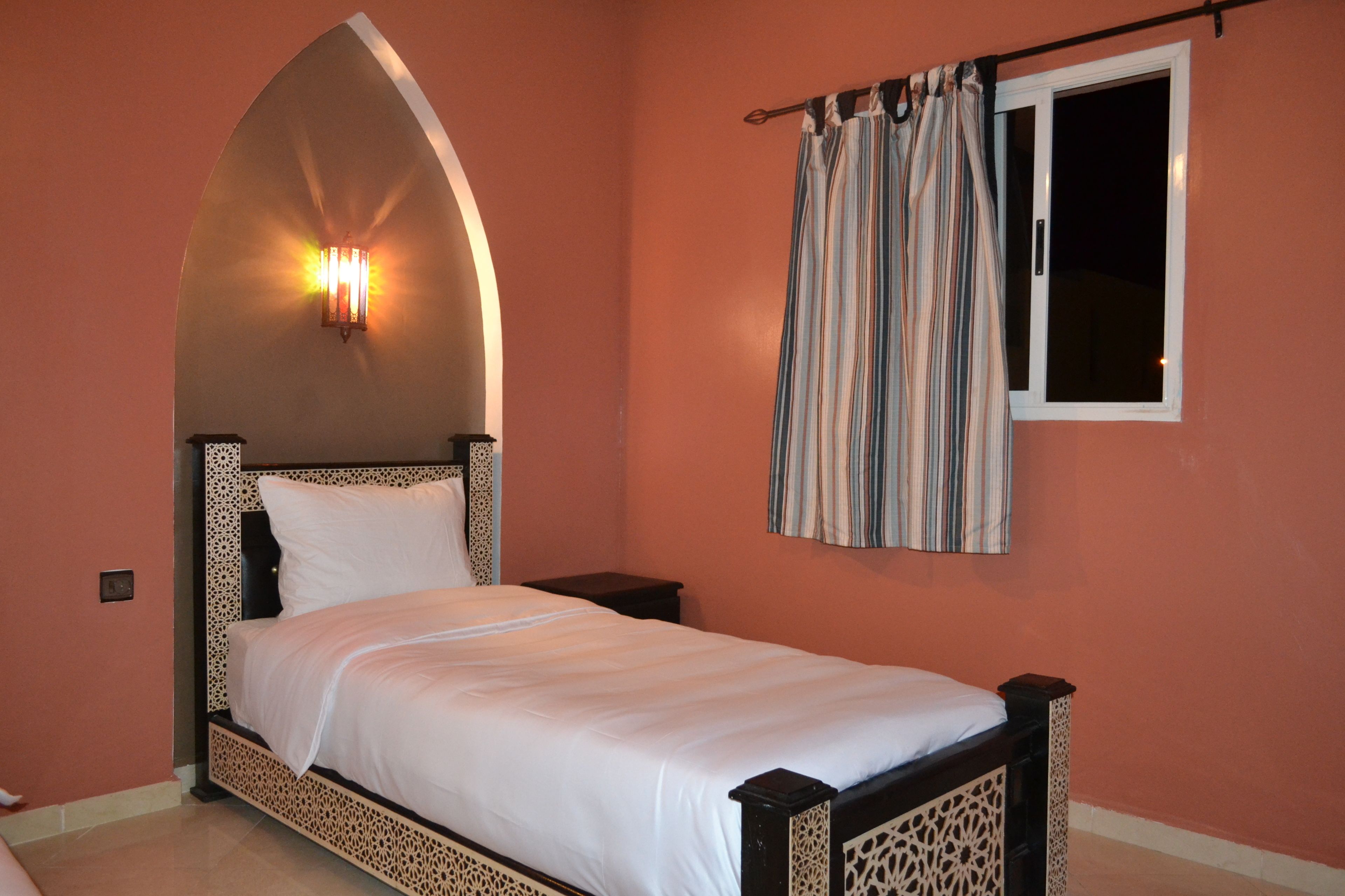 Un dormitorio con cama, mesita de noche, lámpara de pared, ventana con cortinas y puerta.