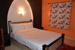 Una cama con colchón y almohadas blancas, cabecera de madera con intrincados grabados, mesita de noche con lámpara y cortina estampada con motivos florales sobre una pared naranja.