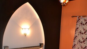 Una cama con una colcha blanca y almohadas, una lámpara fijada a la pared y una cortina con estampado floral.