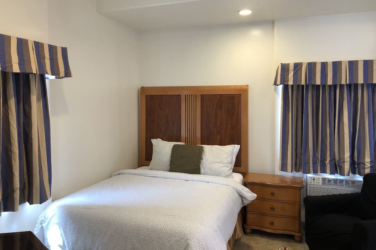 Quarto com cama, cabeceira de madeira, duas mesas de cabeceira, uma cadeira e cortinas.