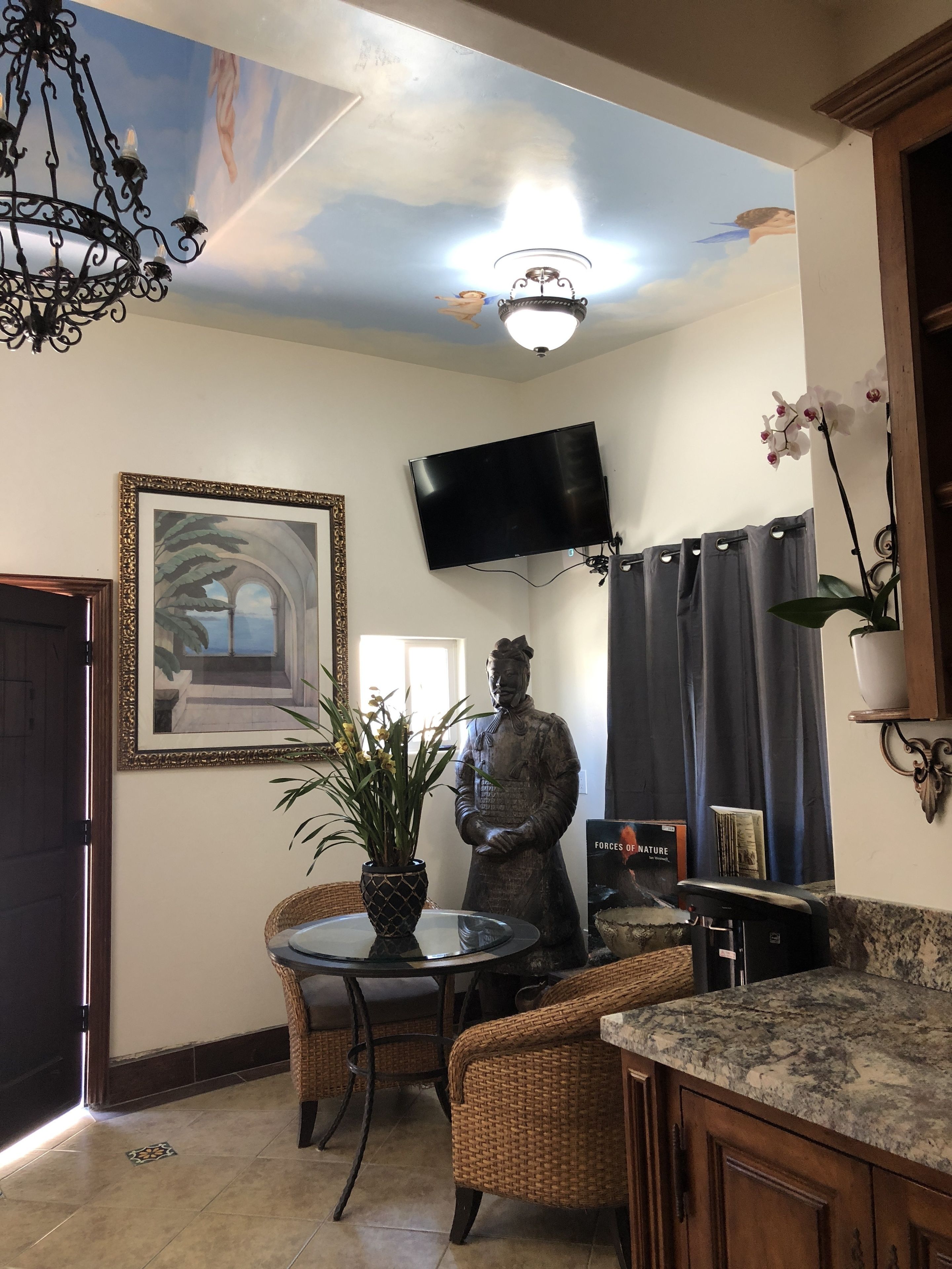 Une pièce dotée d’un ventilateur de plafond, d’un téléviseur à écran plat fixé au mur, d’une statue, d’une plante en pot, de meubles en osier et d’un sol carrelé.