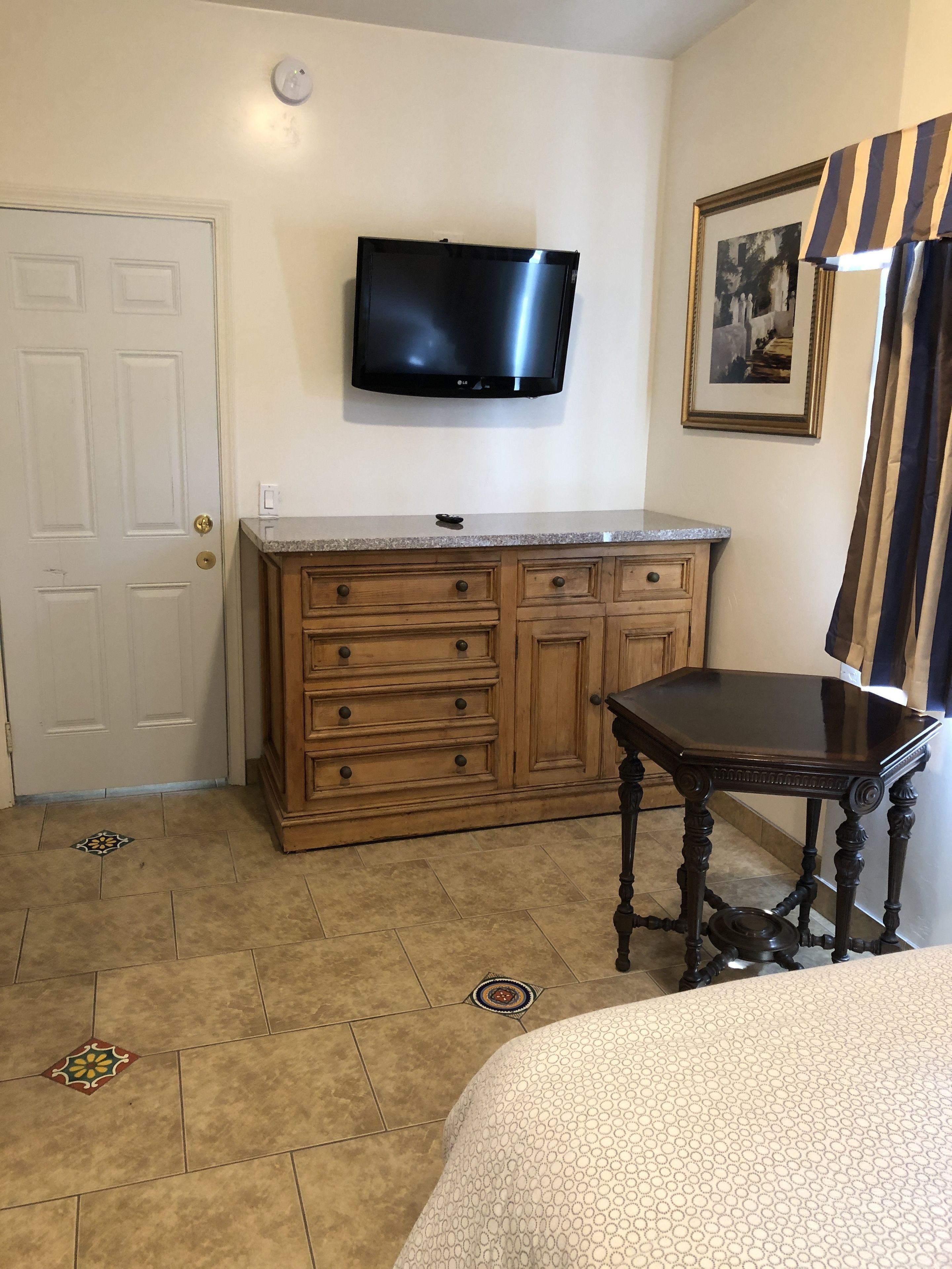 Une pièce comprenant un meuble en bois, un téléviseur à écran plat fixé au-dessus, une table à manger et un lit recouvert d’une couvre-lit à motifs.
