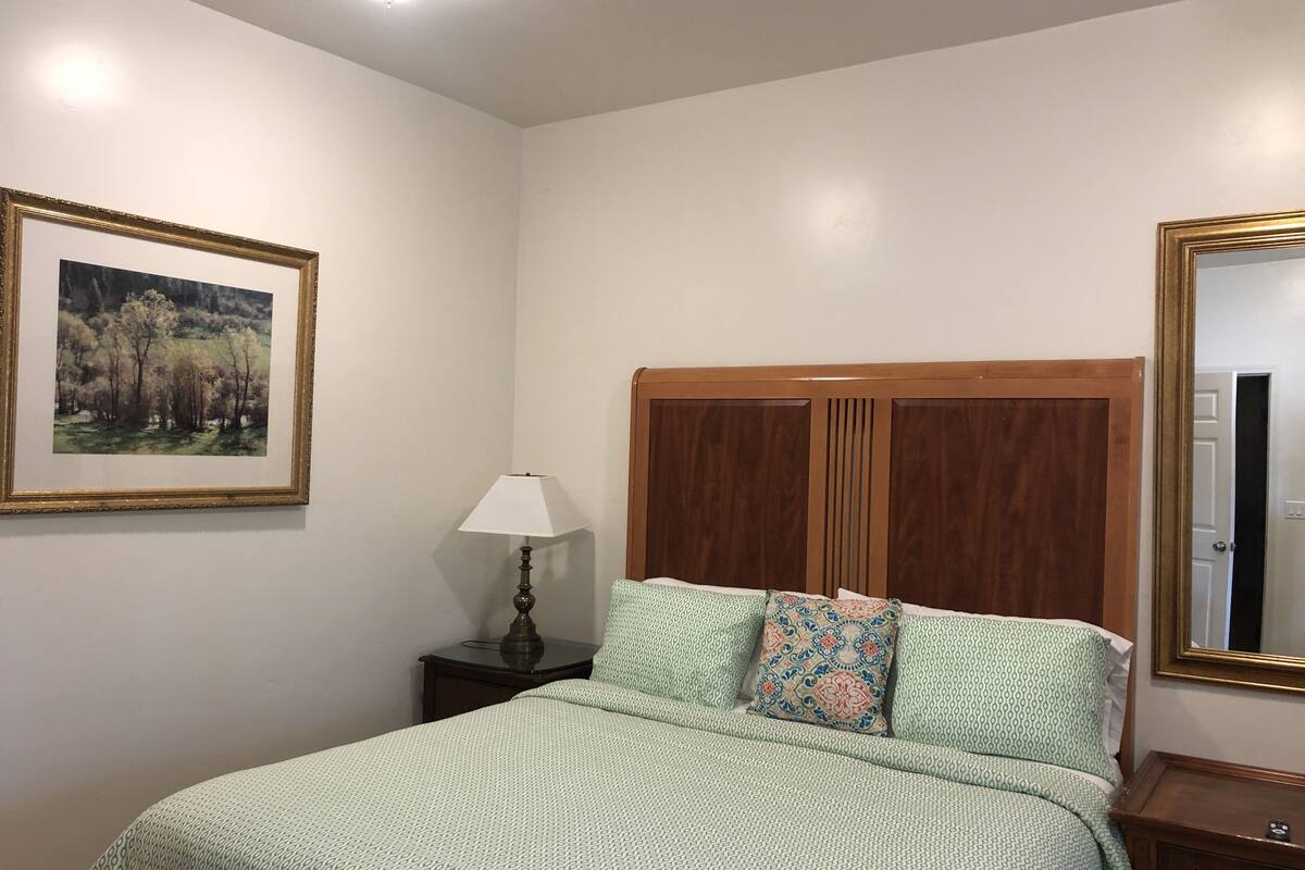 Quarto com cama, mesa de cabeceira, luminária, quadro emoldurado e espelho.