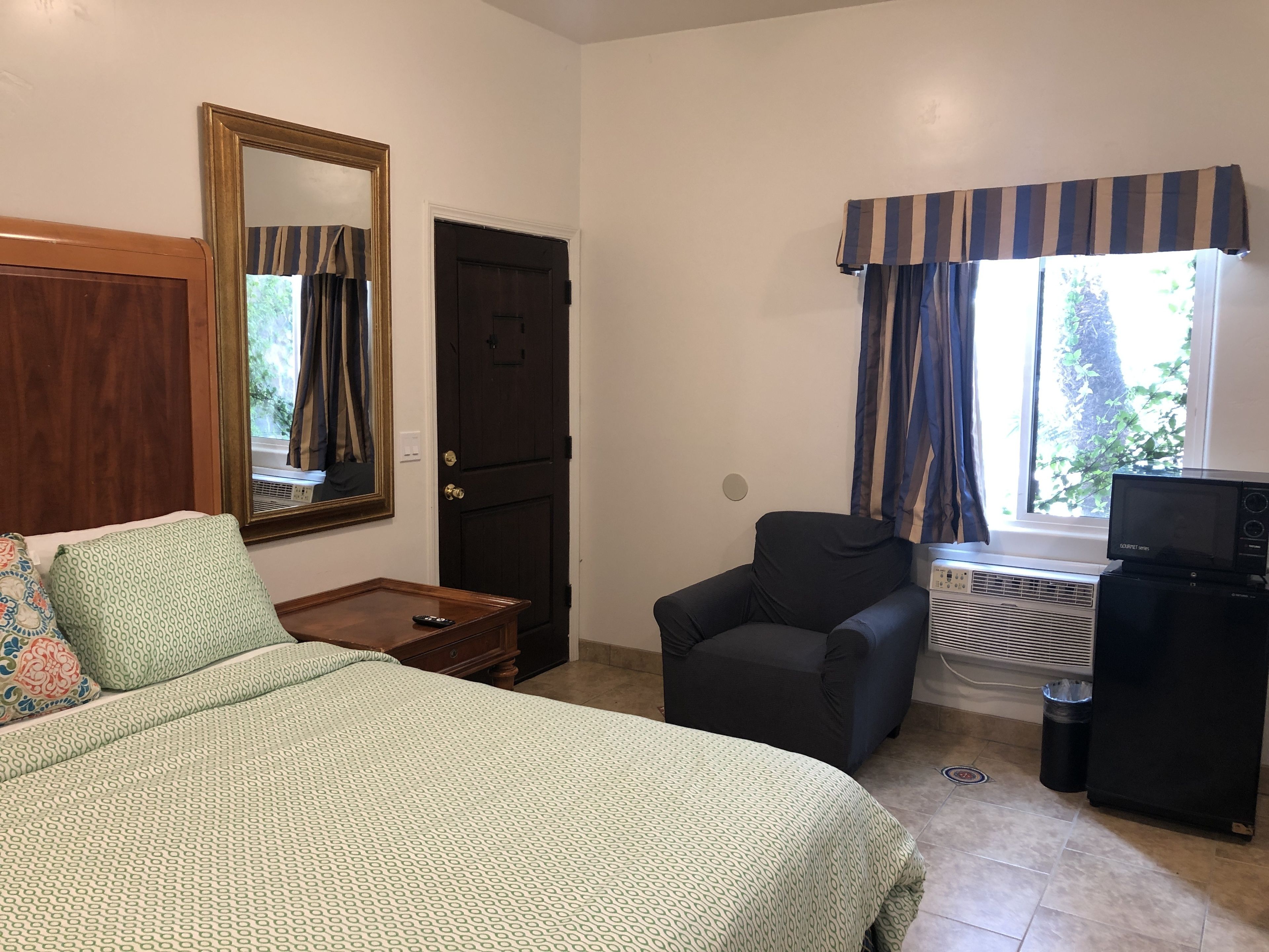 Um quarto com uma cama, uma cadeira, uma escrivaninha com computador, um espelho e uma janela com cortinas.