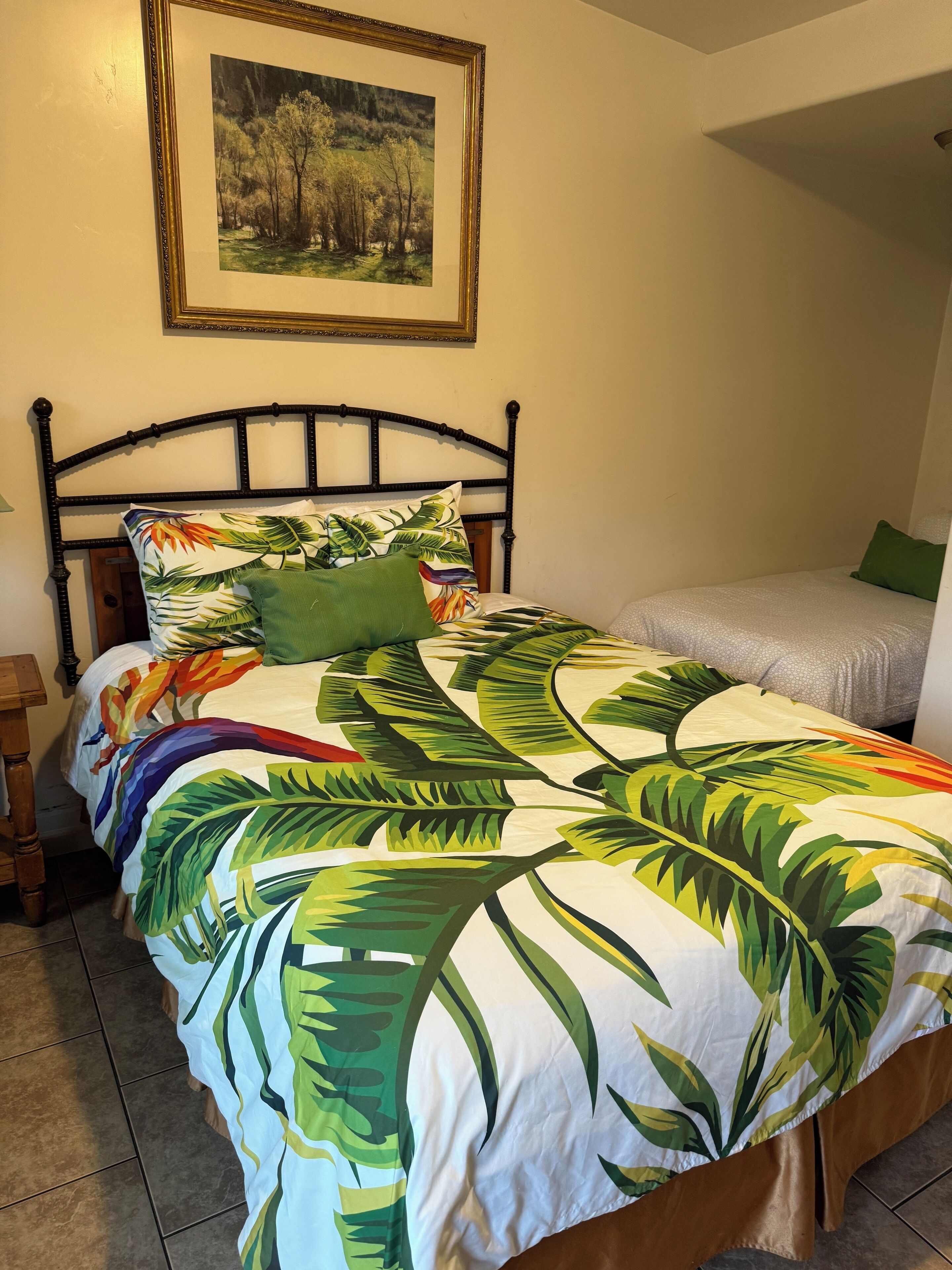 Un letto con una coperta a motivi tropicali, un quadro raffigurante un paesaggio appeso sopra e un comodino con una lampada.