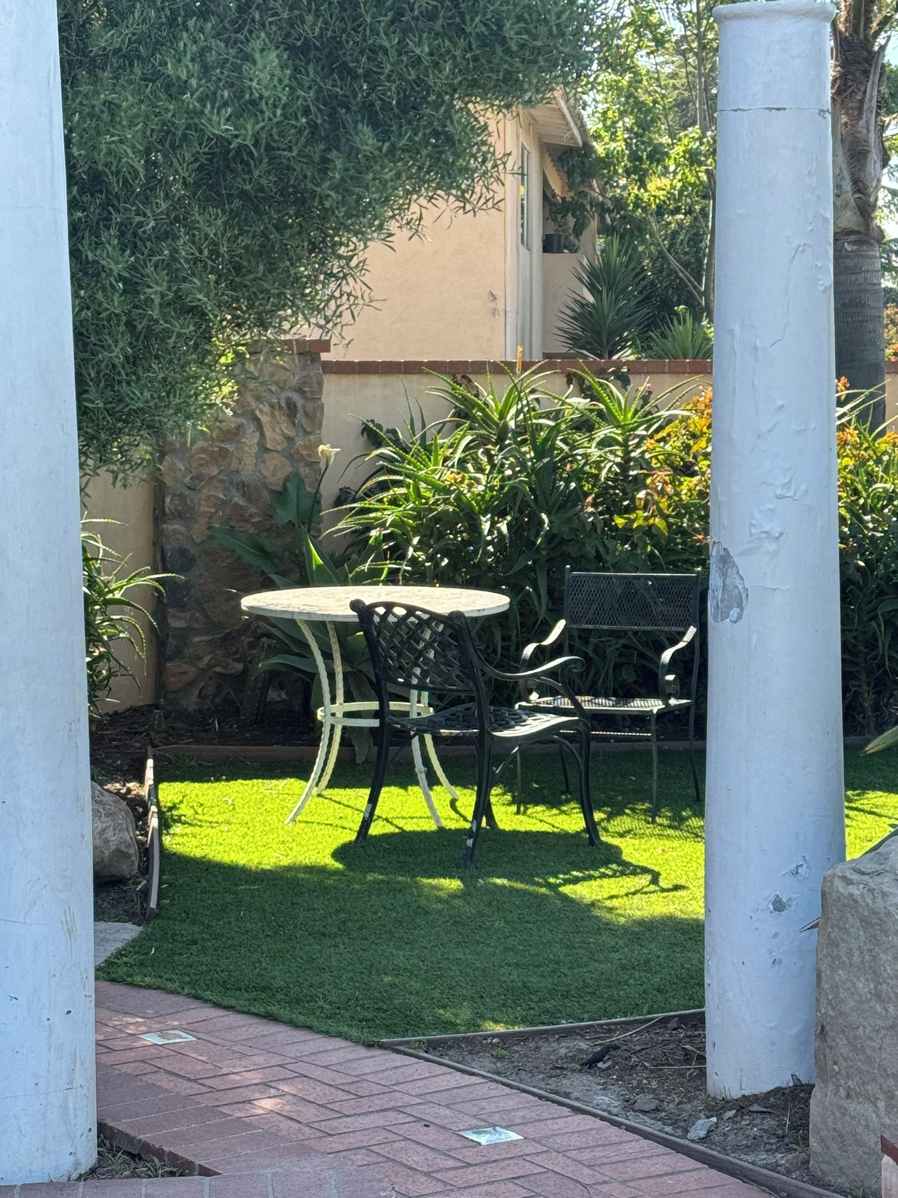 A patio area with a round table and two chairs, surrounded by lush greenery and a stone wall.