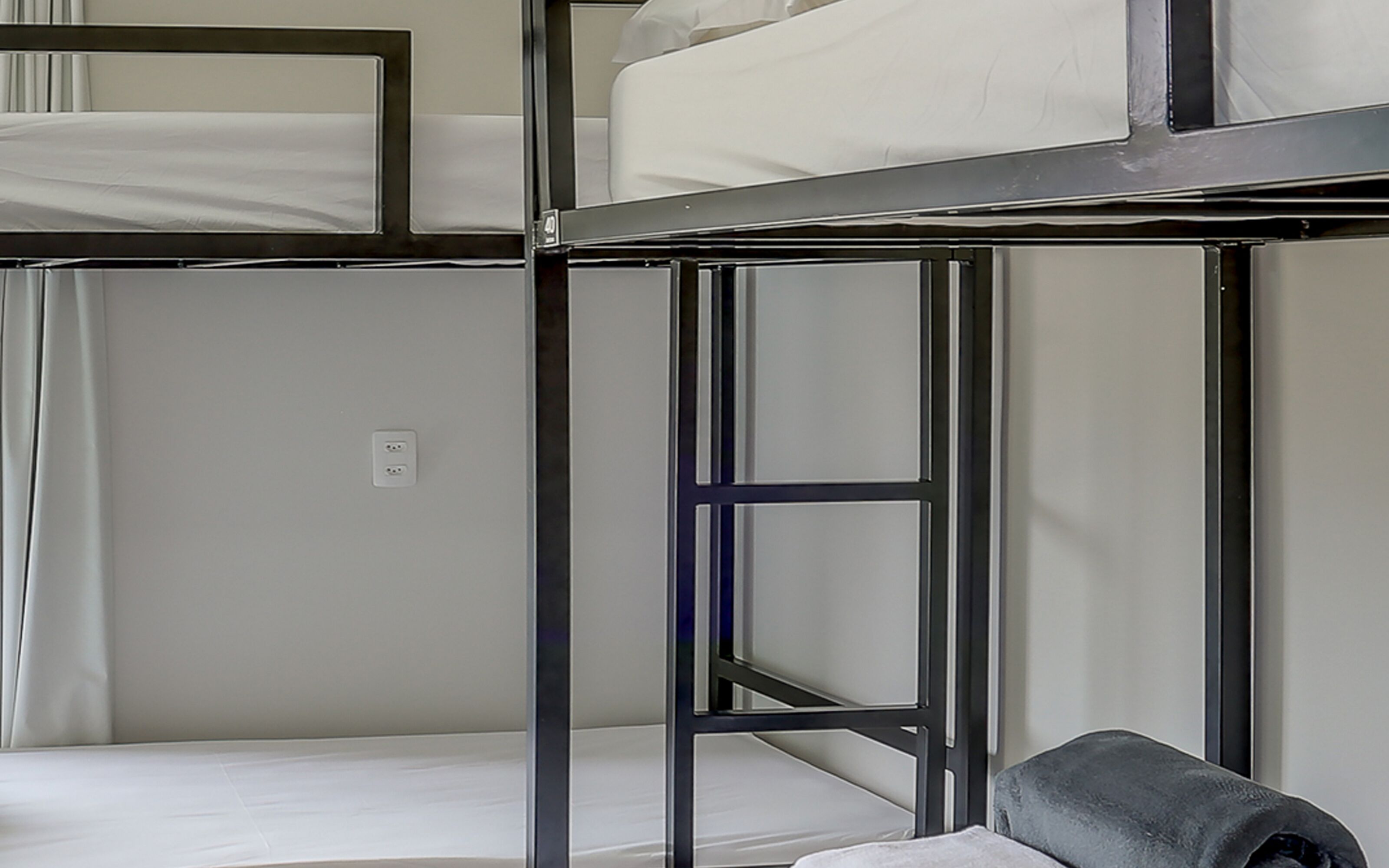 Une chambre avec un lit superposé, une structure en métal, du linge de lit blanc et une prise électrique au mur.
