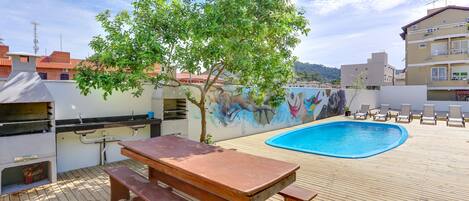 Terraza de madera con una parrilla, mesa de picnic y sillones junto a la piscina.
