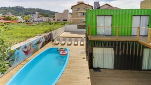 Una piscina con cubierta de madera, un mural y un edificio verde con cortinas blancas.